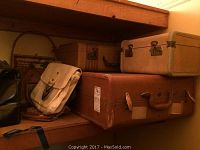 Three hard-side luggage pieces including a large reddish-brown suitcase with handle damage, two beige vintage suitcases, three handbags (white canvas, black, and brown), and a wicker basket on a wooden shelf.