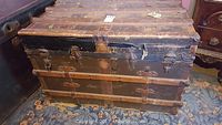 Front view of the vintage wooden steamer trunk showing metal clasps, horizontal wood slats, and worn surface including cracks and repairs