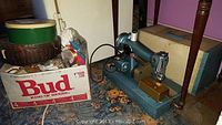 Full view showing the sewing machine on wooden base, carrying case behind, green and cream tin, wicker basket, and a box labeled Bud King of Beers used for storage of sewing supplies