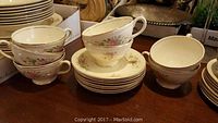 Set of six teacups stacked with matching saucers and a stack of plates with the same floral design and gold rim on a wooden table.