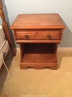 Front view of wood side table showing drawer with two round metal knobs, open lower shelf, and scalloped apron base.