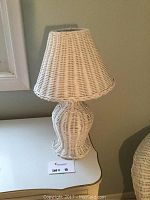 Photo of a white wicker table lamp on a white nightstand, lamp unlit, showing woven wicker base and matching conical wicker lampshade.