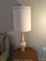 Close-up of one tall table lamp with ribbed base and white drum shade placed on wooden corner table beside couch against a beige wall, no lamp light on.
