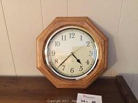 Photo of an octagonal oak trim wall clock hanging on a beige wall, showing clock face and hands clearly.