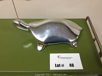 Front and top view of polished aluminum turtle sculpture placed on green surface with numbered lot card.