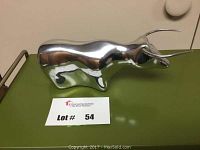 Polished aluminum bull sculpture shown on a surface with lot number card.
