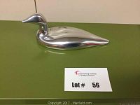 Top-down side view of smooth, polished aluminum loon sculpture on green surface with lot number card.