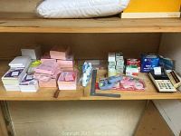 Shelving with multiple stacks of blank memo pads and phone message pads in white and pink tones, several small boxes of staples and paper clips towards the right, tape dispenser with rolls of tape, calculator on the far right, all arranged on a wooden shelf.