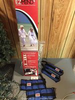 Photo showing EZ Mend red and white boxed hip and knee rehabilitation device, along with York brand weights with blue fabric covers and Velcro fasteners laid out on a gray surface.