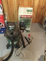 Photo of the Shop-Vac unit with hose, extensions, and attached cords on the floor beside the original packaging box.