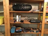 Full view of wooden shelf with RCA portable radio CD player on top shelf with power cord and 4 CDs stacked beside it. Middle shelf contains shoe polish, blue shoe horn, aerosol can, battery charger with cord coiled, and small black tin. Bottom shelf has blue thermos, outdoor electric timers, shoehorn holders, and floor protectors.