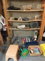 Wooden shelving in workshop with various tools and supplies. Grey space heater, extension cords, plastic wash bins, step stool, caulking materials, and paint roller tray visible.