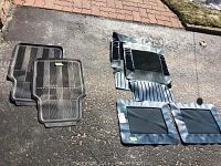 Six black rubber car floor mats arranged on asphalt, showing both 4-piece and 2-piece sets