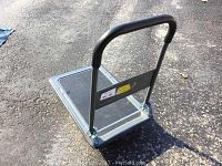 Fold-up cart standing upright on pavement showing metal frame, handle with black grip and platform base.