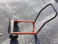 Orange dolly/cart on asphalt with vertical black handle attached at one end, showing metal base plate and two black wheels.