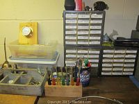 Top view of three metal storage bins with various small hardware items, a wooden caddy holding screwdrivers, and labeled small parts drawers in background