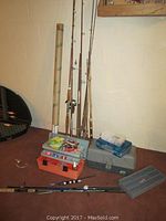 Photo showing multiple fishing poles standing upright against wall with fishing pole tubes, various tackle boxes on floor containing fishing accessories.