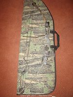 Full view of camo compound bow lying on camo soft case on brown carpet. Shows bow limbs, pulley system, and string.