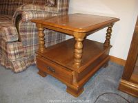 Front view of wooden end table featuring rounded rectangular top, turned spindle legs, lower shelf, and drawer with metal handles.