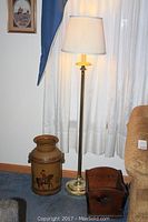 Floor lamp, hand painted milk jug, and wooden magazine holder grouped together near curtain and wall.