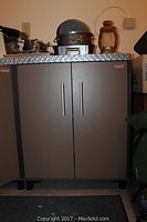 Full front view of the grey Coleman storage cabinet, showing two doors, diamond plate textured top, and items placed on top (not included).