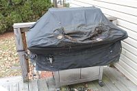 Black grill cover over grill showing tears, dirt and damage on a wooden deck outside next to house.