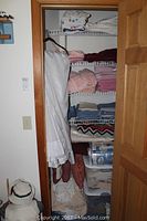 View of linen closet with shelves holding folded towels, linens, sheer drapes, and plastic bins containing table clothes and other linens.