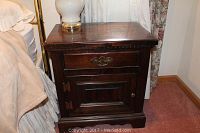 Front view of one wooden end table showing drawer with metal handle and cabinet door with wooden knob. Table has dark wood finish and carved trim details.