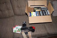 Photo showing Canon AE-1 camera on a couch with zoom lens attached. Two patterned camera straps, multiple film rolls, instruction booklets, and cardboard box filled with DVDs present.