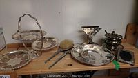 Assorted silver metalware collection including tiered server, serving trays, brush and mirror, small tureen and platter, small boxes and decorative pieces displayed on wooden surface.