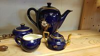 Full view of 4 piece cobalt blue tea set including tall teapot, sugar bowl, creamer, and small teapot with gold accents and floral design