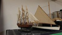 Photo of two wooden model boats seen from side, showing detailed rigging and sails on the three-mast ship and the angled sail on the smaller boat.