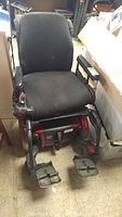 Front view of the motorized wheelchair showing black cushioned seat, armrests, joystick controller, and footrests