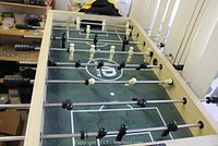 Angled top view of the foosball table showing black and white player figures on metal rods, wooden frame, and playfield markings.