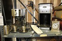 Shelf with Cuisinart coffee maker missing carafe, Cafe Roma espresso maker, metal battery-operated clock, and white Lightning bottle opener.