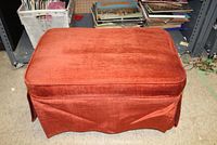 Rust-colored upholstered footstool with skirt and wooden legs shown from the front angle, placed in storage area.