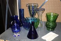 Overview of seven assorted coloured glass items including vases, bowl and pitcher in blue and green colors placed on a shelf.