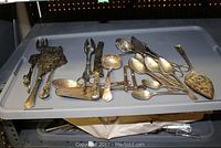 Wide view of assorted silver plated serving utensils including forks, spoons, tongs, and serving knives on a grey plastic tray.