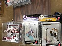 Photo showing three McFarlane NHL hockey player figurines in original plastic packaging on wooden surface: Curtis Joseph Coyotes goalie, a New York Rangers player #11, and Jaromir Jagr #68 Devils player.