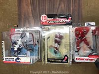 Photo showing three McFarlane NHL hockey figurines in their original packaging displayed side by side. From left to right: Vancouver Canucks player, Steve Yzerman #19 in Detroit Red Wings uniform, Mike Shanahan in red uniform.