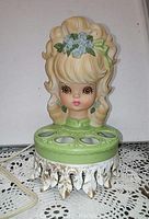 Front view of the ceramic lamp showing the detailed female face and hair with flower decoration, green base with four round lipstick holders and metal stand.
