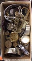 Box containing various wristwatches and small jewelry items piled together. Watches include mix of metal and leather bands, some gold-tone, some silver-tone.