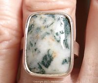 Close-up view of rectangular moss agate stone set in sterling silver ring worn on a finger. Shows stone pattern and setting details.