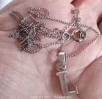 Silver initial 'L' pendant with attached chain shown resting on a palm for scale. Pendant is block style, approximately 7/8 inch tall, and chain is fine linked with spring ring clasp.