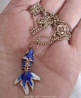 Pendant chain held in hand showing articulated blue fish pendant with gold-colored metal frame and dark blue enamel top surface.