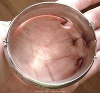 Image of bangle held in hand, showing inner side and overall shape with hinge and safety chain attached.