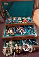 Open jewelry box showing three tiers filled with vintage costume jewelry including earrings, brooches, necklaces, bracelets, and cufflinks on teal velvet lining.