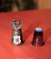 Photo showing two Scottish themed thimbles, one metal with pewter thistle flowers and shield, and one purple glass with Millefiori top.