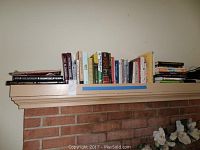 Wide side view of multiple books arranged on a shelf above a fireplace mantel, showing a variety of titles and colors