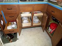 Open kitchen cabinets and drawers showing storage of assorted kitchenware including dishes, cups, bowls, and utensils, with visible signs of wear and age on wooden cabinetry and floor.